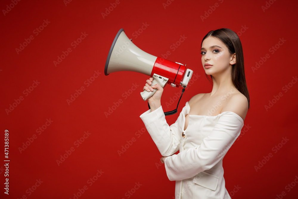 High fashion photo of elegant young model with Megaphone in white suit, pants, trousers top jacket. Beautiful young woman, slim figure. Studio shot. Red background High fashion photo of elegant young model with Megaphone in white suit, pants, trousers top jacket. Beautiful young woman, slim figure. Studio shot. Red background