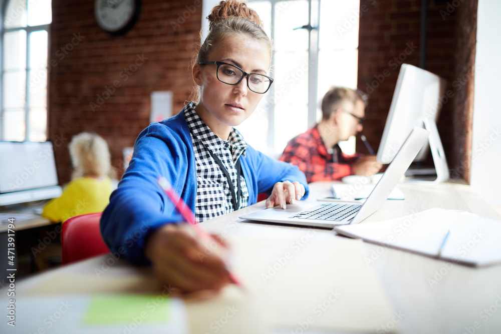 Fototapeta premium Young serious student with laptop making notes while looking for information in the net