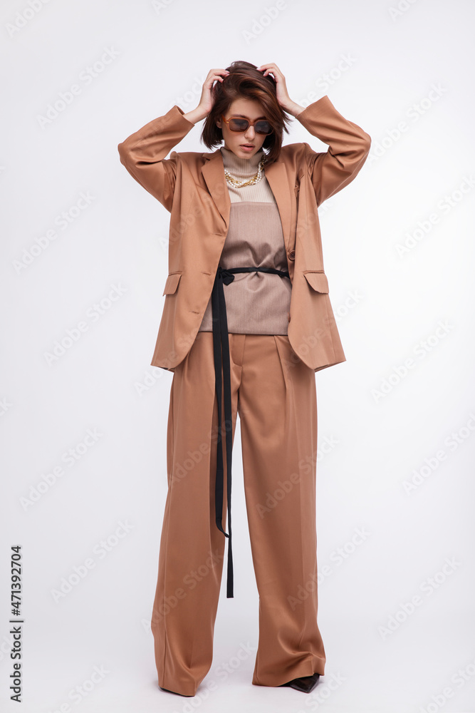 Fashion photo of a beautiful elegant young woman in a pretty oversize brown and beige suit, corset, stylish sunglasses, jacket, pants posing over white background. Bob haircut. Studio Shot. Portrait Fashion photo of a beautiful elegant young woman in a pretty oversize brown and beige suit, corset, stylish sunglasses, jacket, pants posing over white background. Bob haircut. Studio Shot. Portrait