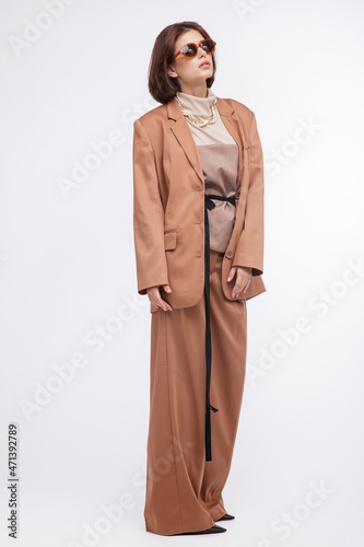 Fashion photo of a beautiful elegant young woman in a pretty oversize brown and beige suit, corset, stylish sunglasses, jacket, pants posing over white background. Bob haircut. Studio Shot. Portrait