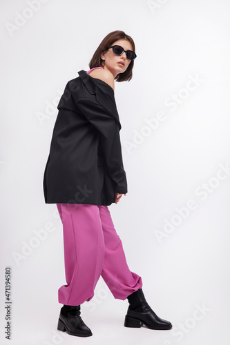 Fashion photo of a beautiful elegant young woman in a pretty oversize black jacket and pink suit, boots, stylish sunglasses posing over white background. Bob haircut. Studio Shot