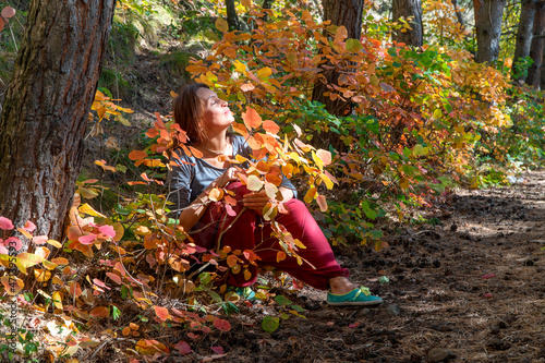 Smiling woman with red Indian pants sitting in the forest under the trees on the path with autumn foliage. Portrait of a happy woman
