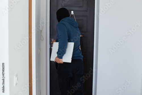 Back view of man robber wearing blue hoodie and black balaclava standing with notebook he stole in hands, leaving place of crime, stole other people's personal belongings.