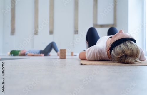 Two women lying on a mat, eyes closed, meditating, during an indoor yoga class.
