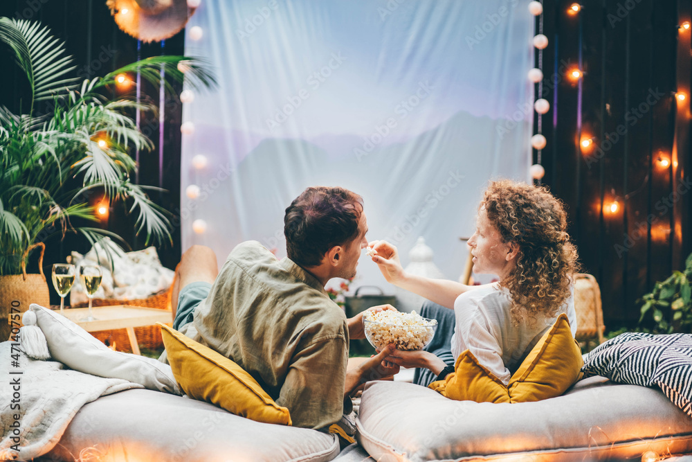 Lovely couple takes delicious popcorn from bowl on pillows in backyard with large fabric as screen on wall at romantic date close backside view.