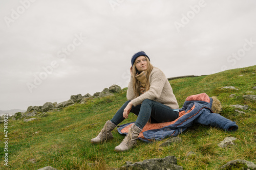 Young beautiful woman in knitted sweater and hat is sitting on her coat on ground. Cloudy day. Scandinavian nature.