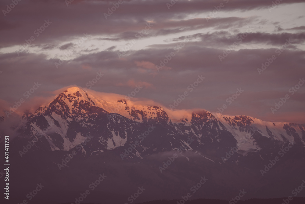 Scenic mountain landscape with great snowy mountain range lit by dawn sun among low clouds. Awesome alpine scenery with high mountain ridge at sunset or at sunrise. Big glacier on top in vintage color