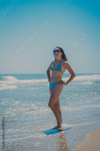 A young woman posing in the summer on the beach. A girl n in a blue swimsuit and fashionable sun glasses stands on the sand. Ocean or sea on the background. A female rests and takes sunbath.