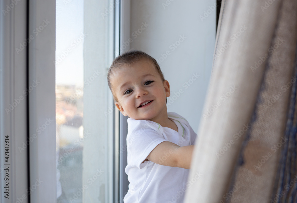 little boy smiling standing on the windowsill by the window behind the curtain little boy smiling standing on the windowsill by the window behind the curtain