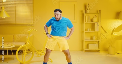 Slow motion of funny handsome young man in blue t-shirt doing aerobics with silly movement at home with yellow interior. Bearded male model in sportswear having a workout with funny expression