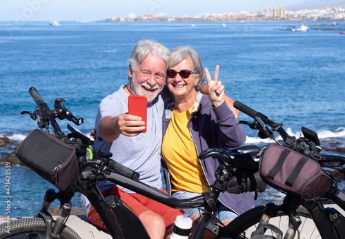 Joyful beautiful senior couple using phone in outdoor excursion near their bicycles, horizon on the sea. Active retiree enjoying healthy lifestyle