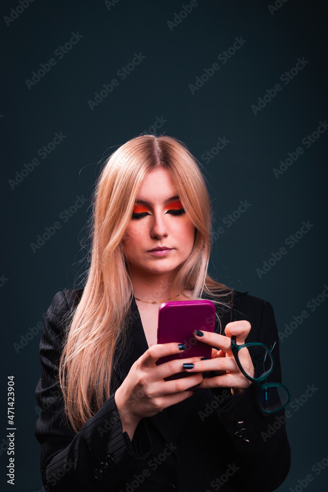 Young caucasian business woman using a mobile phone wearing a black blazer and glasses in a photography studio with a blue dark background. Young caucasian business woman using a mobile phone wearing a black blazer and glasses in a photography studio with a blue dark background.