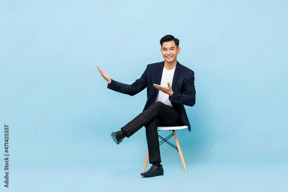 Smiling young Asian man sitting cross legged with hands open to empty space aside in light blue isolated background Smiling young Asian man sitting cross legged with hands open to empty space aside in light blue isolated background