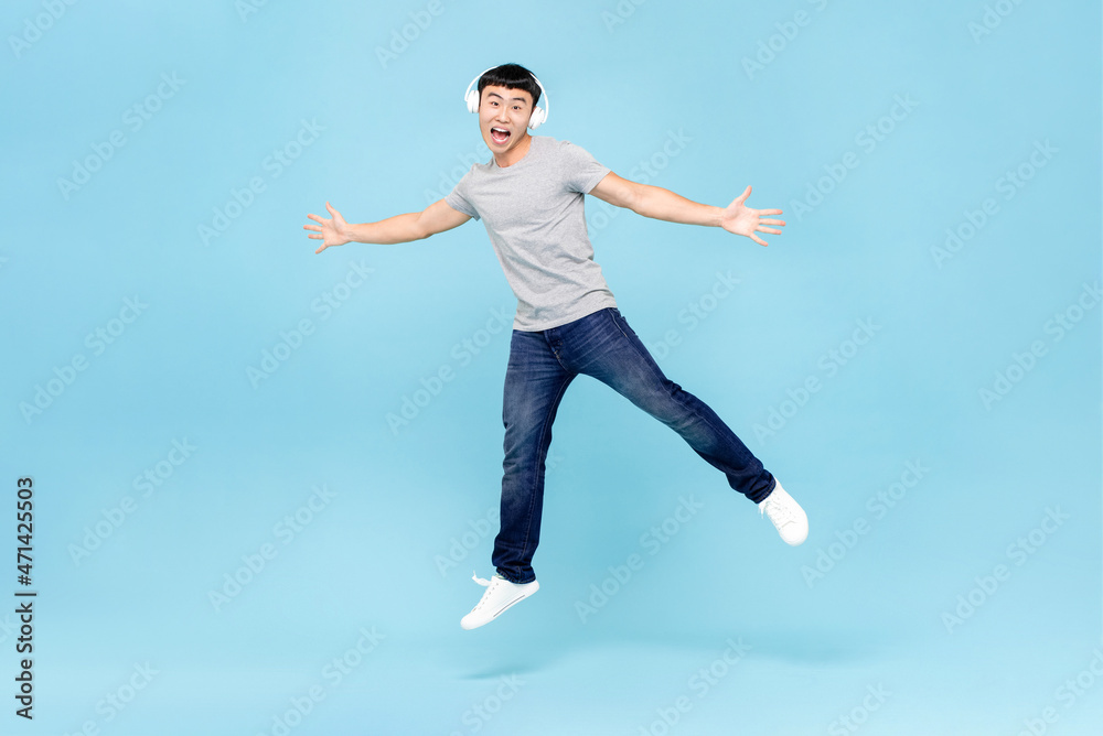 Energetic young Asian man wearing headphones listening to music and jumping on light blue isolated background