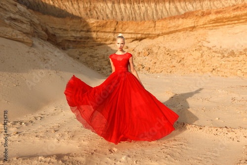 Full length image of a young woman in red waving dress with flying fabric on the background of golden sands.