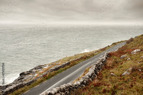 Small narrow asphalt road by ocean. Rough terrain. Burren area, west of Ireland. Low cloudy sky. Stone fences by a road. Wild Atlantic Way route. Travel and transportation.