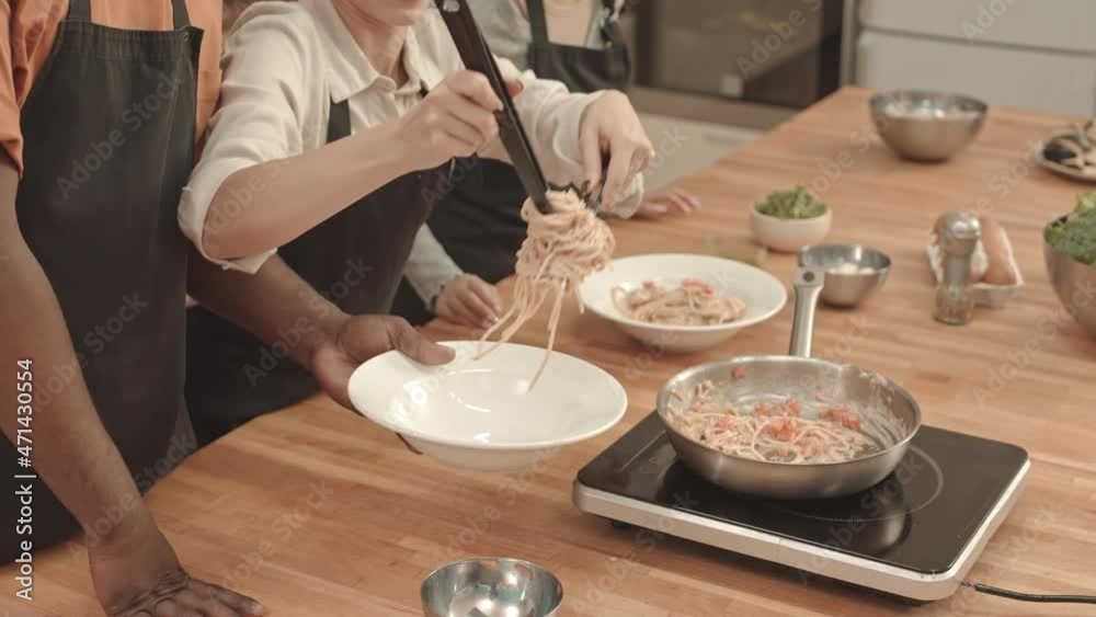 High angle of young Caucasian woman using tongs and ladle for putting pasta from pan into plate that cropped African American man holding, Asian culinary student watching High angle of young Caucasian woman using tongs and ladle for putting pasta from pan into plate that cropped African American man holding, Asian culinary student watching