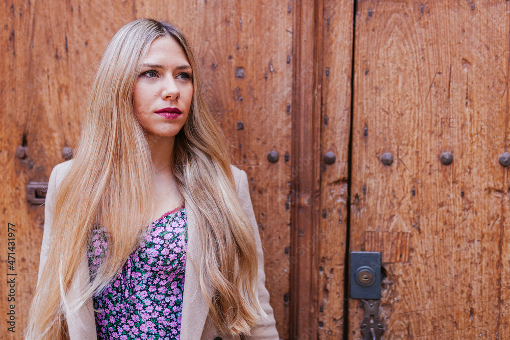 Fototapeta premium Blond latin woman leans against old door in the street. Stylish argentinian woman with scar on cheek with winter clothes.