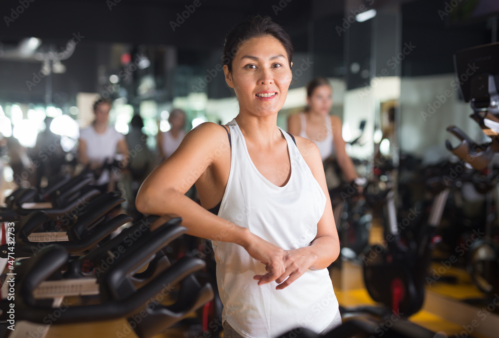 Smiling young adult woman standing near stationary bike before cardio workout in gym