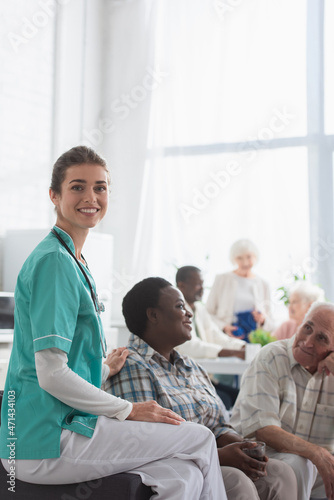 Young nurse smiling at camera near multiethnic patients in nursing home