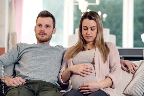 Fototapeta Naklejka Na Ścianę i Meble -  Young pregnant happy couple sitting on couch husband stroking baby belly of blond wife