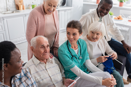 Senior man holding digital tablet near nurse and interracial friends in nursing home