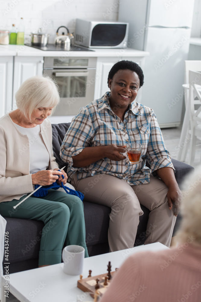 Smiling african american woman with tea sitting near friend knitting and chess in nursing home Smiling african american woman with tea sitting near friend knitting and chess in nursing home