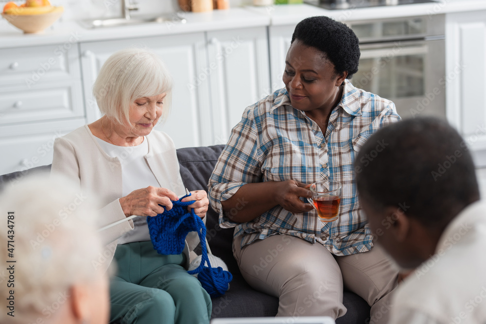 Fototapeta premium Senior woman knitting near african american friend with tea in nursing home