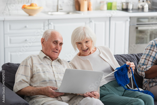 Senior man holding laptop near cheerful friend with yarn in nursing home