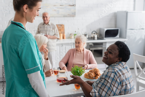 Smiling african american woman talking to nurse near tea and croissants in nursing home
