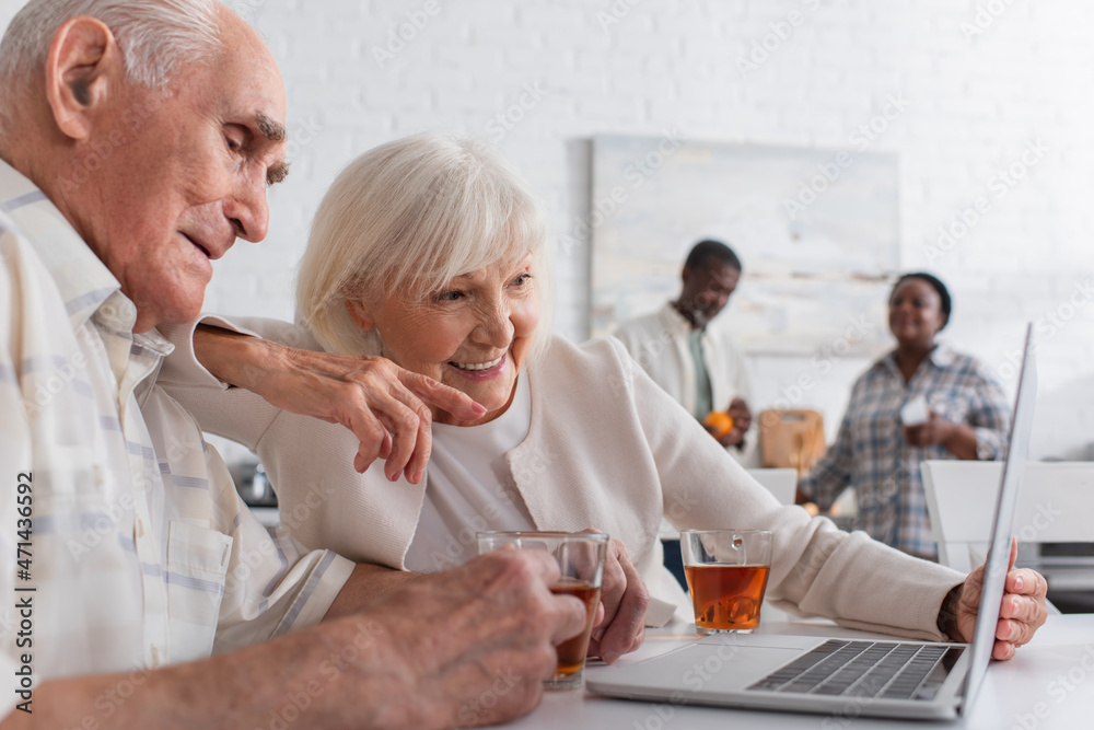 Obraz premium Positive elderly woman pointing at laptop near friend with cup of tea in nursing home