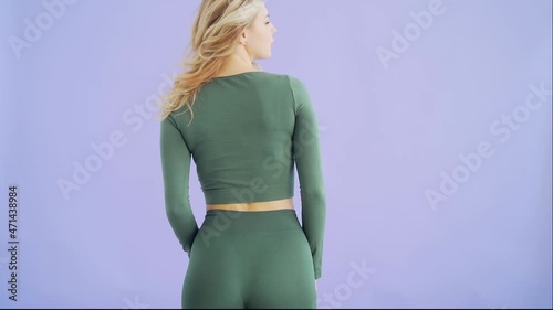 Portrait young attractive fitness woman athlete in tracksuit posing looking at camera. Beautiful female blonde girl in green sportswear on purple or blue studio background. Coach with slender figure