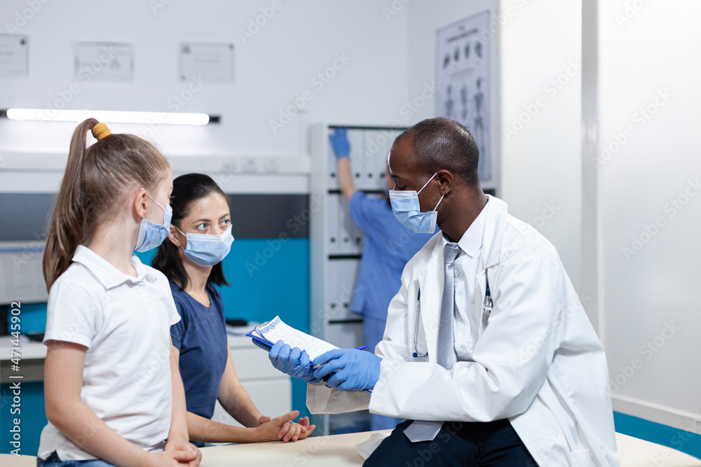 Fototapeta premium African american pediatrician doctor analyzing sickness symptoms expertise discussing healthcare treatment with parent during clinical appointment in hospital office. Medical team with face mask