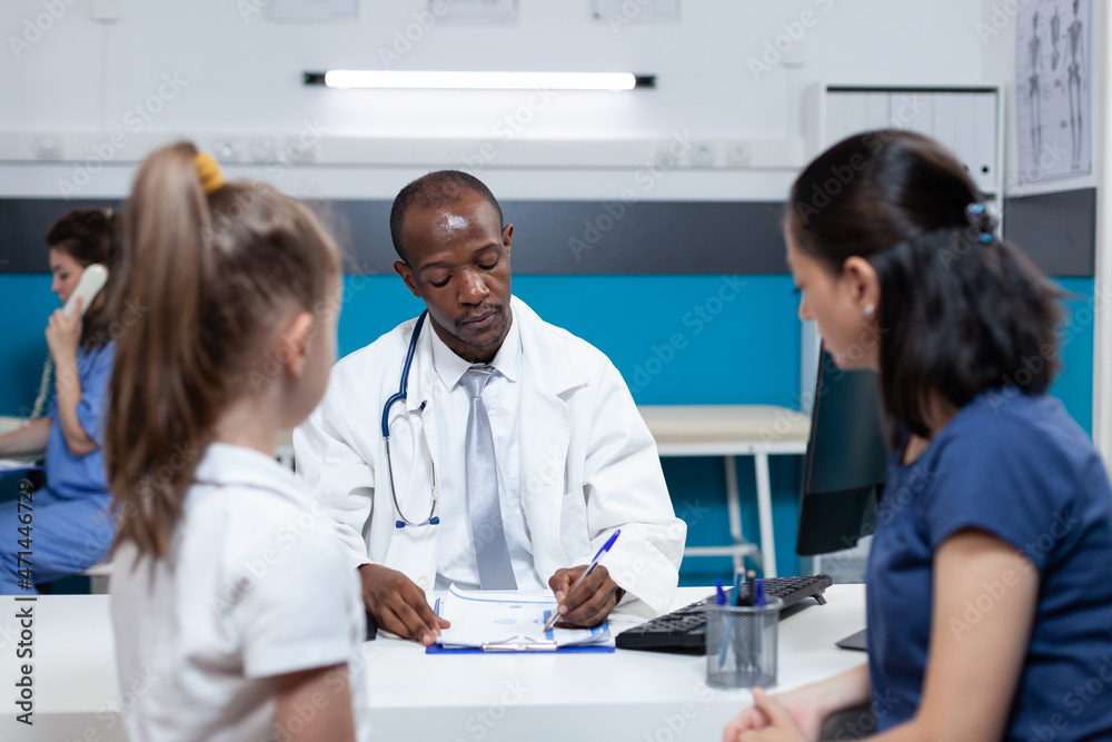 Fototapeta premium African american pediatrician doctor writing sickness symptoms on clipboard discussing medical treatment with family during clinical examination in hospital office. Health care treatment