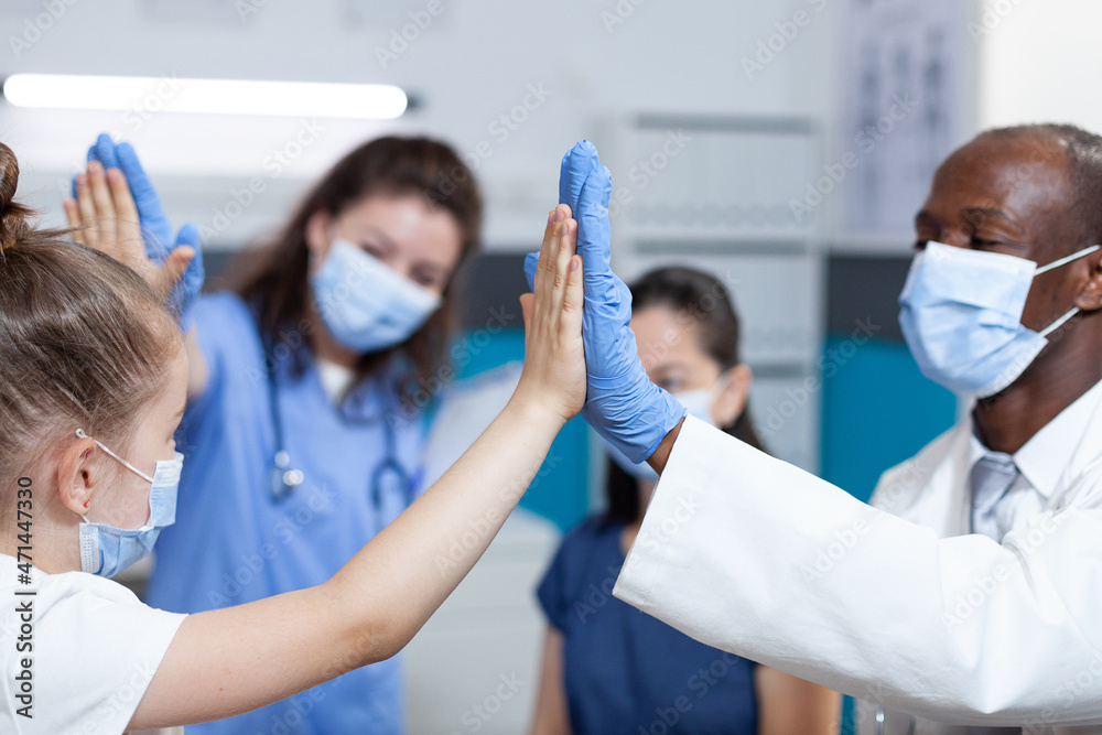 Young girl patient with protection face mask against covid19 giving high five to medical team during clinical consultation in hospital office. Pediatric doctor discussing healthcare treatment Young girl patient with protection face mask against covid19 giving high five to medical team during clinical consultation in hospital office. Pediatric doctor discussing healthcare treatment