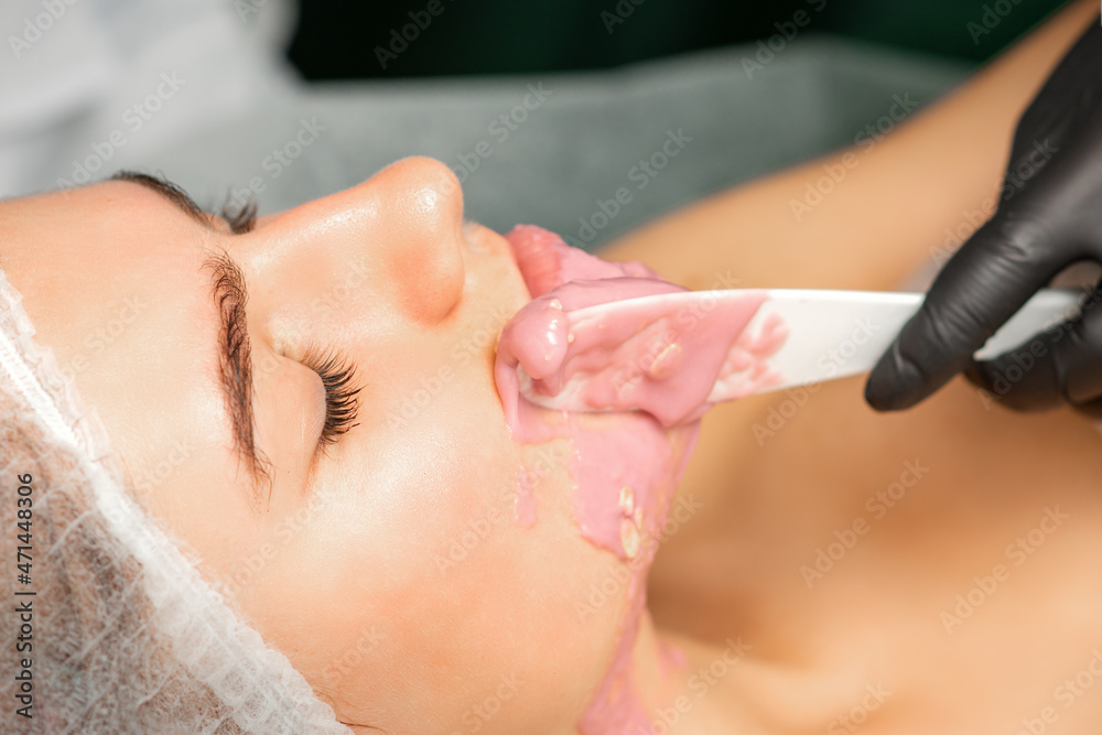 Beautician applying alginate peel-off powder facial mask with the spatula in a spa