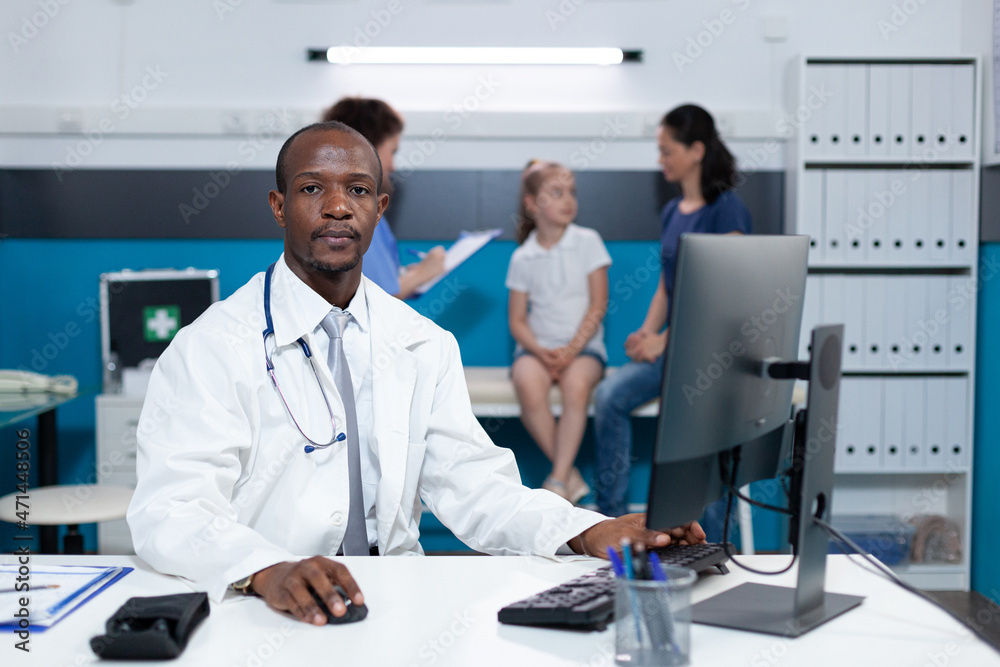 Portrait of african american practitioner doctor sitting at desk in hospital office analyzing sickness expertise on computer during clinical appointment. Pediatric man typing illness symptoms