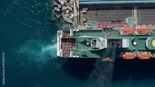 Top down aerial view freight and passenger ferry ship reaching destination in port dock. Carrying cars and people by sea ocean. Exhaust emissions from working engines. White foam in deep blue water