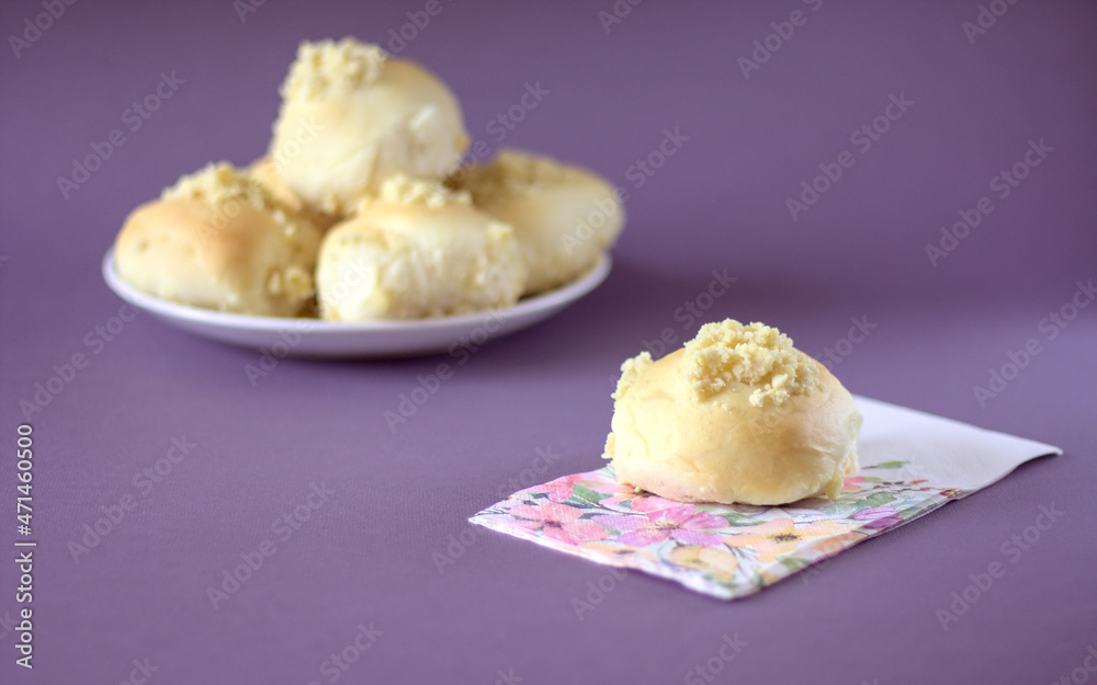 Homemade yeast roll (bun) with crumble on a flowery napkin. Pleasant gray - violet background, in the distance a plate with buns. Copy space.