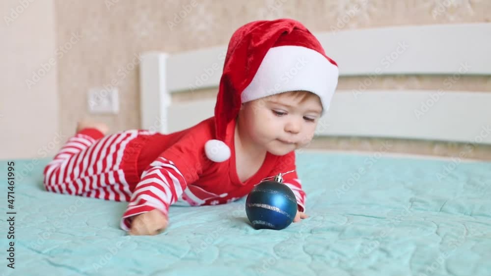 A one-year-old baby lying on tummy wearing a red and white Christmas Santa hat and suit and playing with blue Christmas ball. Merry X-mas and happy new year, infants, childhood, holidays concept A one-year-old baby lying on tummy wearing a red and white Christmas Santa hat and suit and playing with blue Christmas ball. Merry X-mas and happy new year, infants, childhood, holidays concept