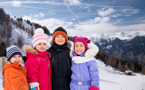 childhood, friendship and season concept - group of happy little kids in winter clothes outdoors over snow-covered mountains or alps on background