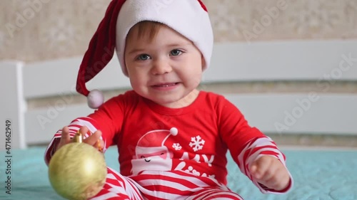 Little Caucasian baby in Santa hat playing with golden Christmas balls indoors. Christmas at home and people concept - charming baby wearing red and white Christmas suit
