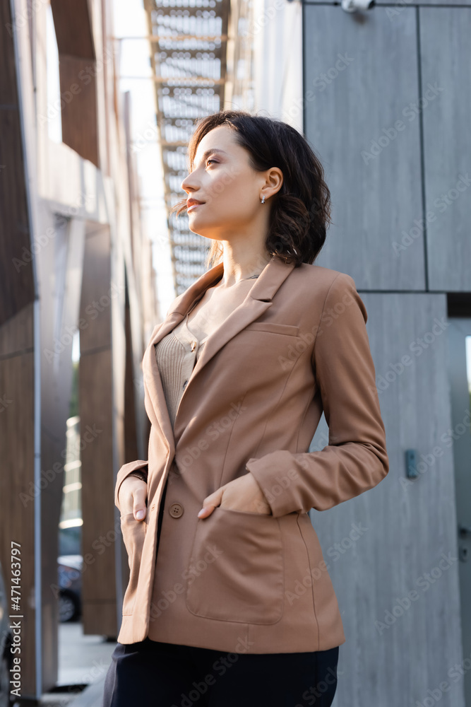 young woman looking away while standing with hands in pockets of beige blazer outdoors