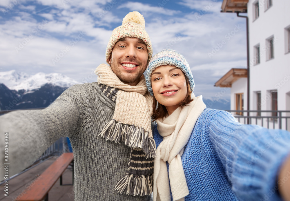 travel, tourism and winter holidays concept - happy couple in knitted hats and scarves taking selfie over mountains and ski resort background