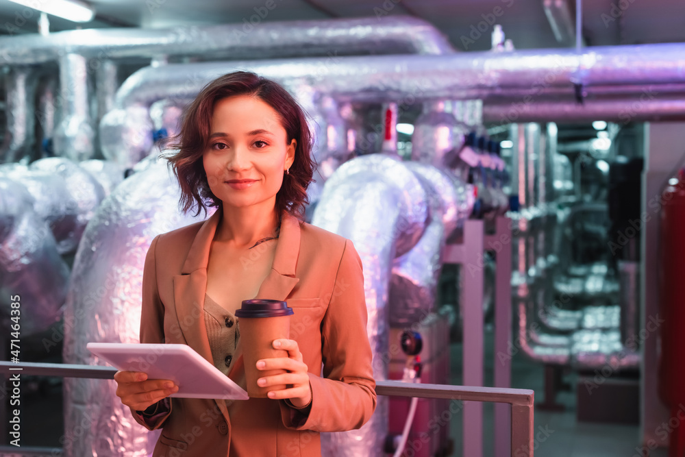 young engineer with digital tablet and coffee to go smiling at camera in technical room of data center young engineer with digital tablet and coffee to go smiling at camera in technical room of data center