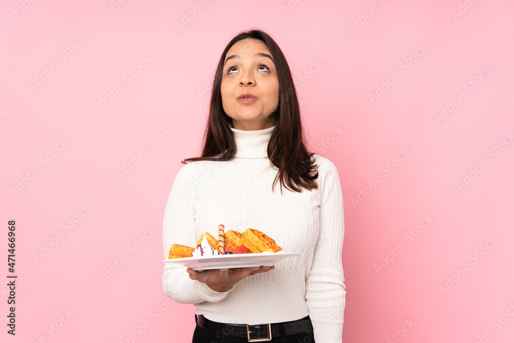 Young brunette woman holding waffles over isolated pink background and looking up Young brunette woman holding waffles over isolated pink background and looking up
