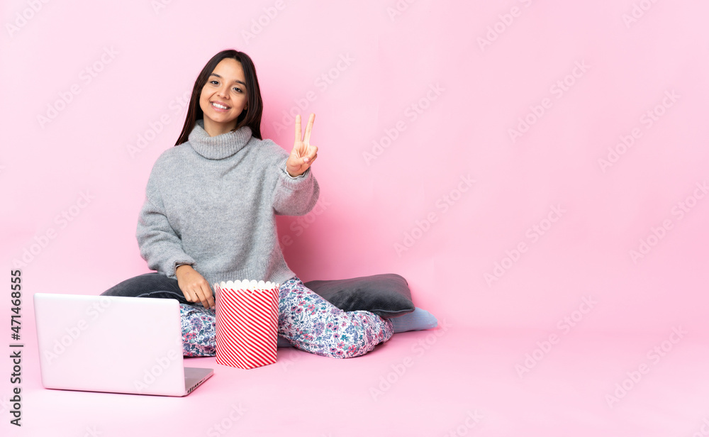 Young mixed race woman eating popcorn while watching a movie on the laptop smiling and showing victory sign Young mixed race woman eating popcorn while watching a movie on the laptop smiling and showing victory sign