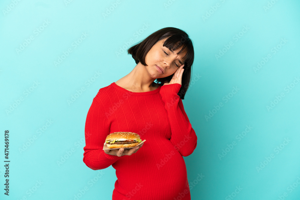 Young pregnant woman holding a burger over isolated background with headache