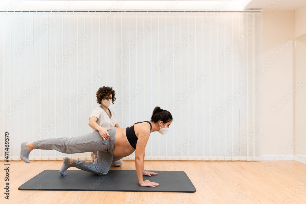 Obraz premium A pregnant woman with face mask kneeling on mat doing pilates on a mat with help of physiotherapist with copy space