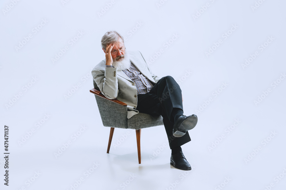 Portrait of senior bearded man in retro style, fashion of 70s, 80s years sitting in chair isolated on gray studio background Portrait of senior bearded man in retro style, fashion of 70s, 80s years sitting in chair isolated on gray studio background